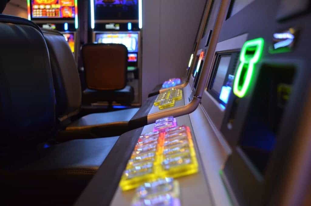 Hintereinander mehrere stationäre Spielautomaten aufgereiht, von denen im Dunkeln nur leuchtende Knöpfe und Teile der Displays zu sehen sind.